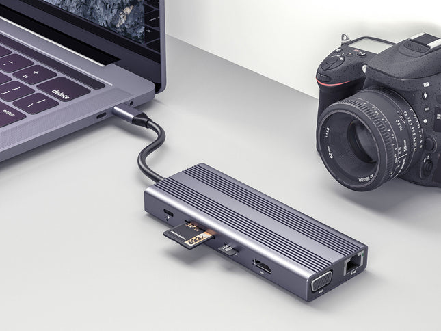 A portable HDMI laptop adapter in a dark metallic gray finish, shown connected to a silver laptop on the left and a black DSLR camera on the right, set against a clean light gray background.