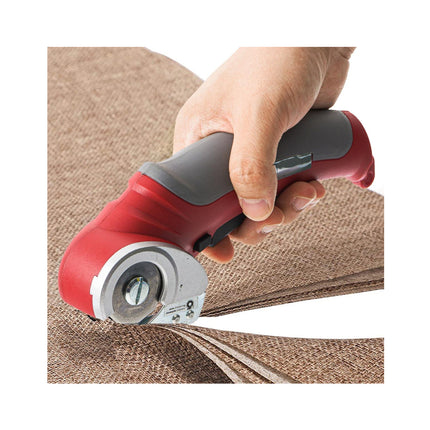 A close-up of a person's hand holding a red and gray rotary cutter over brown burlap fabric, cutting through a stack of similar fabrics against a white background.