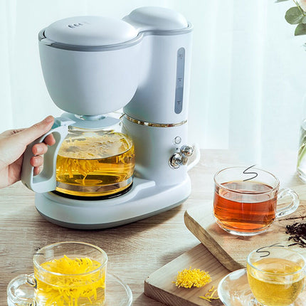 A light gray electric drip coffee maker with a glass carafe filled with brewed coffee, shown on a wooden table with cups of tea and a hand holding the carafe handle.