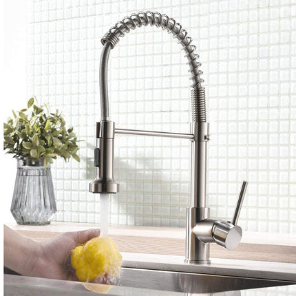 A modern pull-down kitchen faucet with a flexible spring coil handle and a single lever handle, shown installed in a sink with water running. A hand holding a yellow sponge is visible under the faucet, and a small vase of green leaves sits on the counter behind it.