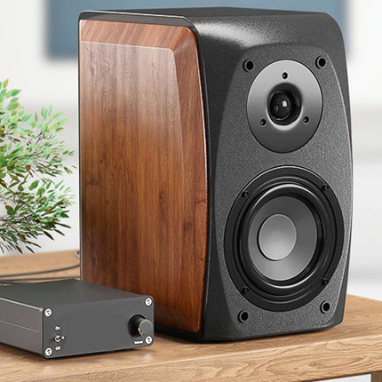 A pair of bookshelf speakers with a dark gray textured front and wood-grain side panels, shown alongside a small black amplifier on a wooden table with a blurred indoor background and a potted plant.