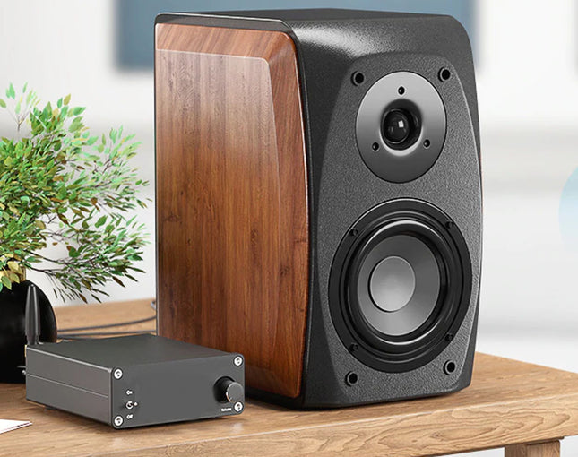 A pair of bookshelf speakers with a dark gray textured front and wood-grain side panels, shown alongside a small black amplifier on a wooden table with a blurred indoor background and a potted plant.