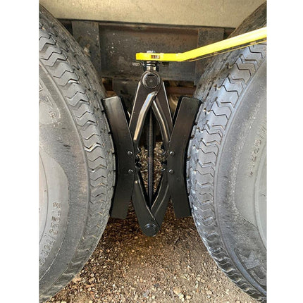 A black metal tire lifter jack positioned between two vehicle tires, with a yellow handle extending upward, photographed outdoors on gravel.