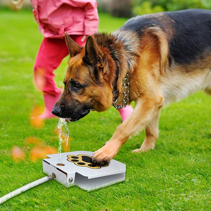 A German Shepherd dog drinks water from a silver automatic dog fountain on a green grass lawn, with a child in a pink outfit standing in the background.