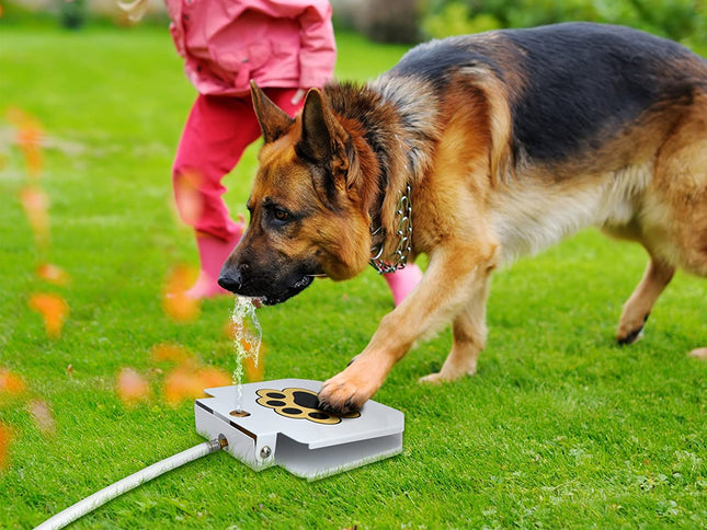 A German Shepherd dog drinks water from a silver automatic dog fountain on a green grass lawn, with a child in a pink outfit standing in the background.