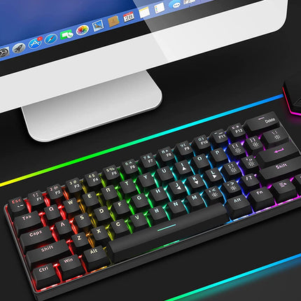 A black mechanical gaming keyboard with RGB rainbow backlighting, shown on a dark desk next to a computer monitor and a glowing RGB desk lamp.