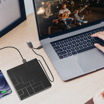 A black rectangular wireless power bank sits on a wooden desk, connected to a smartphone on the left and a laptop on the right. A person's hands are visible using the laptop, and a coffee cup is in the foreground, indicating a lifestyle workspace setting.