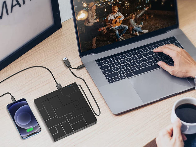 A black rectangular wireless power bank sits on a wooden desk, connected to a smartphone on the left and a laptop on the right. A person's hands are visible using the laptop, and a coffee cup is in the foreground, indicating a lifestyle workspace setting.