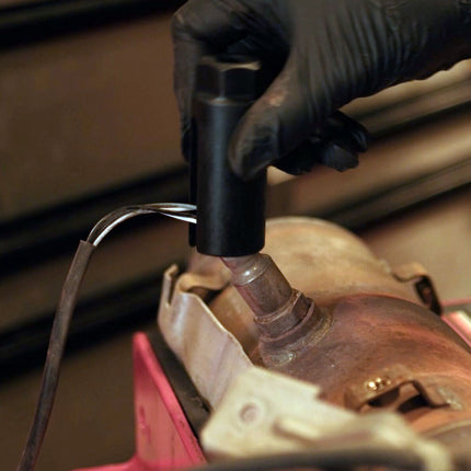 A close-up action shot of a person wearing a black glove using a black handheld tool to remove or adjust a bolt on a rusty metal part, with a pink workbench surface visible in the foreground.