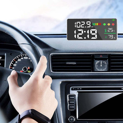 A digital car dashboard display screen mounted above the air vents, showing the time, speed, temperature, and battery status. A person's hand is resting on the steering wheel inside a car cabin.