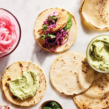 Tortillas with avocado spread, pickled onions, and other toppings on a marble surface.