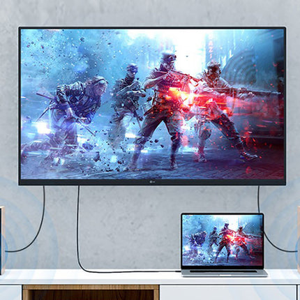 A large wall-mounted TV displaying a blue and red video game scene, with a laptop in front of it and two blue speakers on either side, set against a light gray textured wall.