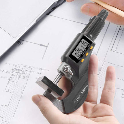 A digital micrometer caliper held in a person's hand over white architectural blueprints, with a pencil in another hand and part of a laptop visible.
