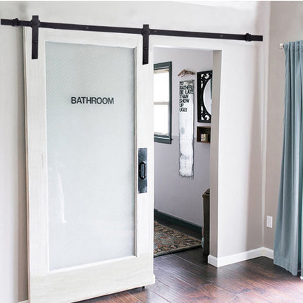 A white-framed sliding barn door with a frosted glass panel labeled 'BATHROOM,' installed on a black metal track in a home interior with wood flooring and a blue curtain.