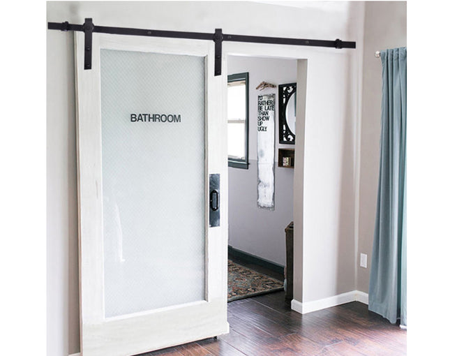 A white-framed sliding barn door with a frosted glass panel labeled 'BATHROOM,' installed on a black metal track in a home interior with wood flooring and a blue curtain.