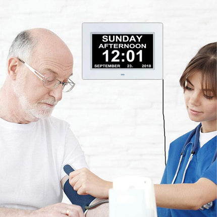 A digital wall clock with a black screen and white frame mounted on a white brick wall above a scene of an elderly man having his blood pressure checked by a female healthcare worker.