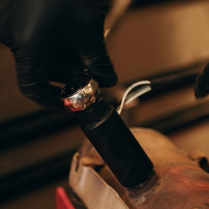 A close-up shot of a person wearing black gloves adjusting a metallic silver and black handle on a leather item, set against a dark, warm-toned workshop background.