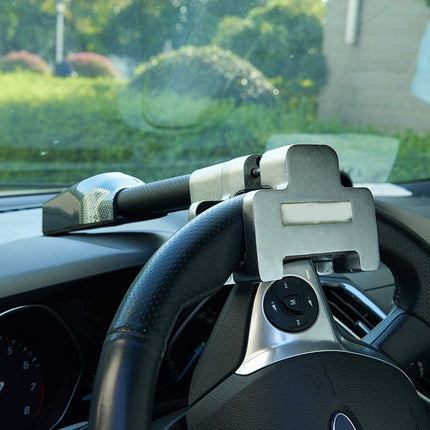 A silver and black car steering wheel accessory mounted on the steering wheel, shown inside a vehicle with a blurred outdoor background visible through the windshield.