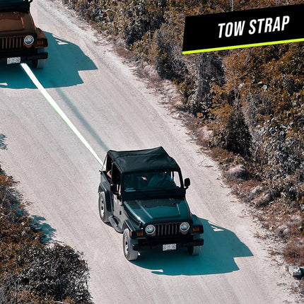 A black tow strap with a bright yellow accent line is shown in a lifestyle outdoor setting, extending from a dark green Jeep to another vehicle on a dirt road. A black banner in the top right corner reads 'TOW STRAP'.