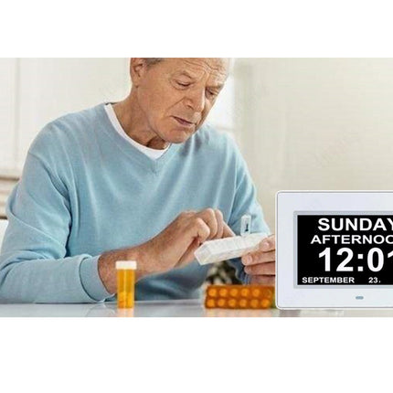 A digital wall clock with a white frame and black screen displaying 'SUNDAY AFTERNOON 12:01 SEPTEMBER 23, 2018' shown next to an older man sitting at a table holding pills.
