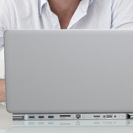 A silver aluminum laptop shown from the back, with ports along the bottom edge, positioned in front of a person wearing a white and blue striped button-down shirt.