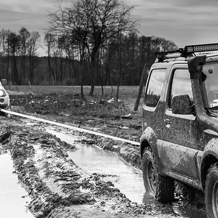 A black and white outdoor scene showing two off-road SUVs on a muddy track, with one vehicle being towed by a rope from another vehicle. Trees and an overcast sky are visible in the background.