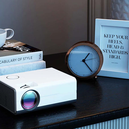 White projector on a wooden surface with books, a clock, and a framed quote in the background.