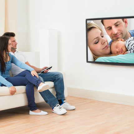 Family sitting on a couch watching a video of a couple with a baby on a TV screen.