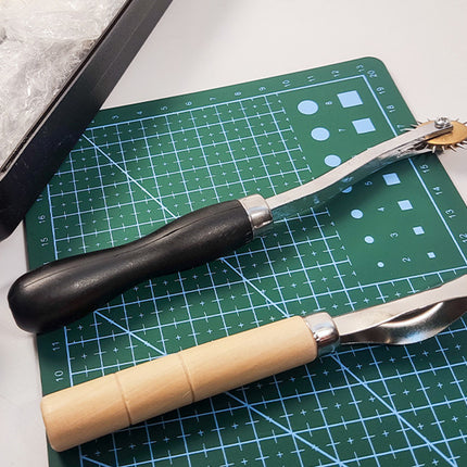 Two leatherworking tools with black and light wood handles resting on a green cutting mat, with part of a black case visible at the top left.
