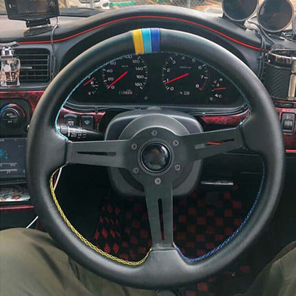 A close-up of a black racing-style steering wheel with a blue, yellow, and white stripe at the top, blue contrast stitching, and a black center hub. The wheel is shown installed inside a car cabin with a red-and-black checkered floor mat and dashboard instruments visible.