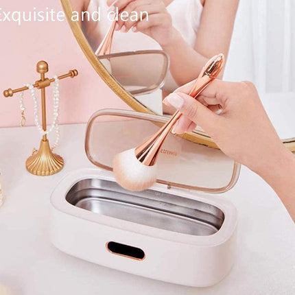 A hand holds a rose gold makeup brush over an open white and rose gold electric makeup cleaner with a clear lid, set on a white vanity table next to a gold jewelry stand and mirror.