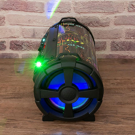 A cylindrical portable speaker with a black body, colorful circuit board graphics, blue LED light glowing from the front speaker grille, and green laser/projector lights emitting beams, set on a wooden surface against a brick wall background.