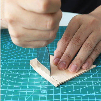 Close-up of a person's hands using a metal scoring knife to cut a light beige leather strip on a green cutting mat with grid lines.