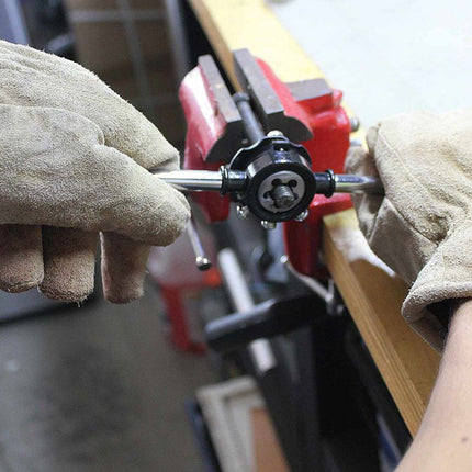 Close-up of a person wearing heavy-duty tan leather welding gloves using a tool on a black and red clamp fixture mounted on a wooden workbench.