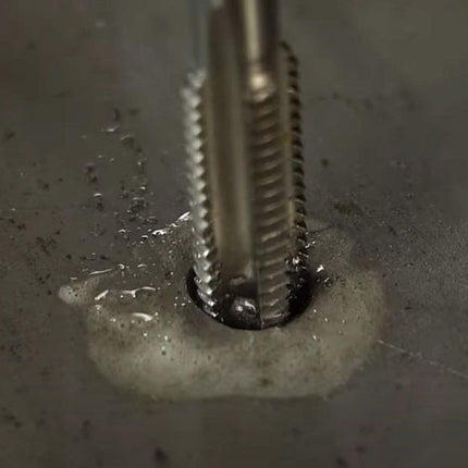 A close-up of a metal threading tool bit or drill bit actively creating a thread on a dark metallic surface with visible coolant or oil mist.