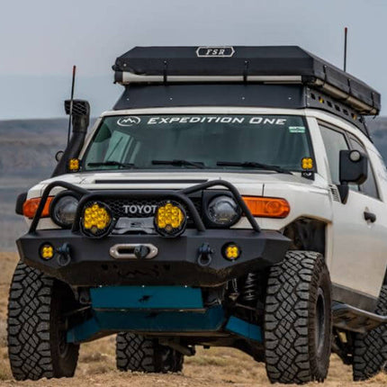 A white Toyota FJ Cruiser off-road vehicle with a black roof box, front bumper guard, and bright yellow auxiliary lights, parked in a dry desert landscape.