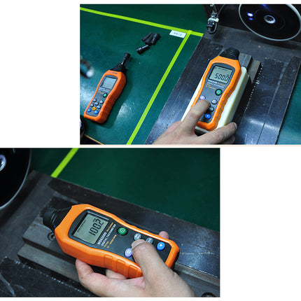 Two handheld orange-and-black digital measuring devices with LCD screens are shown being used on a dark metal surface over a green workbench. A hand is visible holding the devices, and one device lies nearby on the bench.