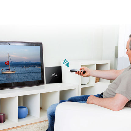 A man sits on a white sofa in a bright living room, holding a remote control pointed toward a television on a white shelf. The TV displays a sailboat scene, and a small black device labeled 'Xbox' sits beside it on the shelf.