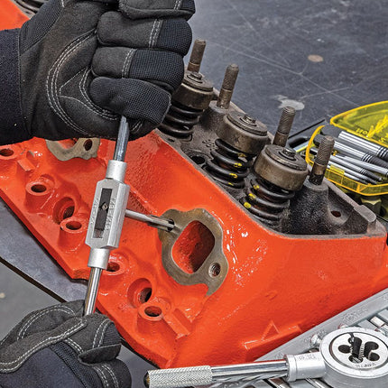 A close-up of a person wearing black gloves using a silver metal tap tool on an orange engine block, with engine components and a yellow tool case visible in the background.