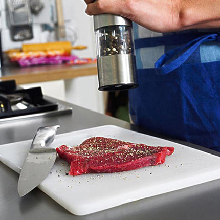 Person seasoning a steak on a cutting board with a pepper grinder in a kitchen setting.
