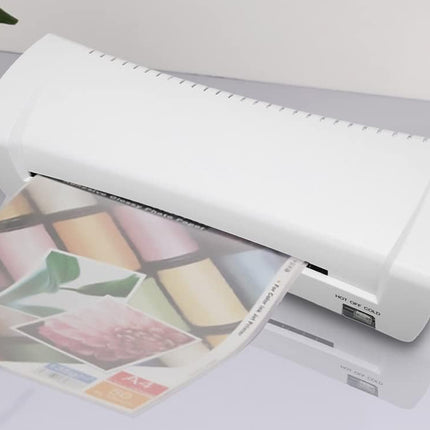 A white desktop document laminator with a glossy finish, shown in the process of laminating a printed sheet of glossy paper. The product is photographed on a light gray reflective surface with a small green plant leaf visible in the top-left corner.