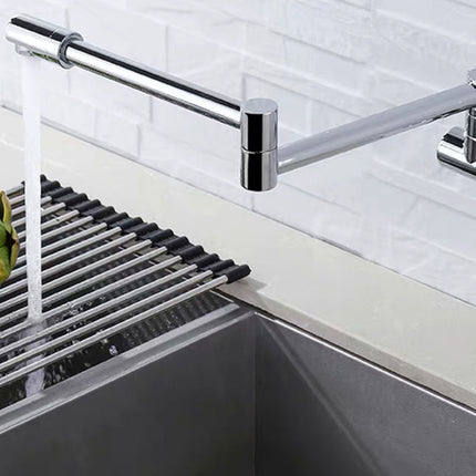 A modern chrome wall-mounted kitchen faucet with a long horizontal spout, shown with water running into a stainless steel sink. A black slotted drain basket is placed inside the sink, and a green artichoke is partially visible on the left.