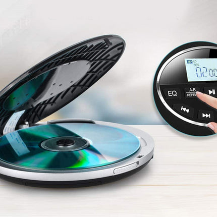 A compact CD player with a disc inserted, shown open on a light wood surface against a soft gray background. A circular inset on the right shows a close-up of the control panel with a finger pressing buttons.