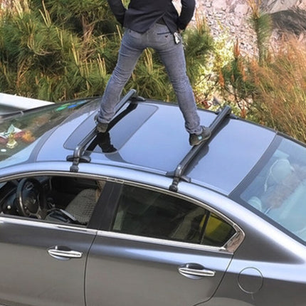 A person standing on top of a silver sedan, demonstrating a black roof rack system installed on the vehicle. The photo is taken outdoors with grass and rocks in the background.