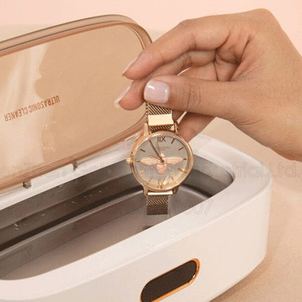 A gold-tone watch with a mesh strap is being held by a hand above an open white ultrasonic cleaner with a rose-gold interior.